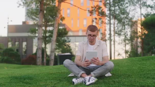 現代の大学公園眼鏡をかけた若い学生の男は、草の上に座っている間に大学のプロジェクトを作るために電子タブレットを使用して — ストック動画