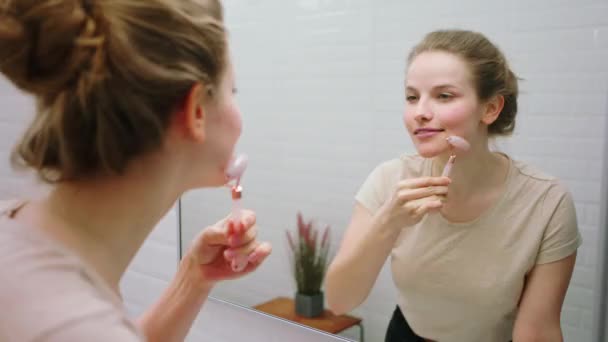 W świetlistej łazience piękna kobieta pozywa wałek do twarzy, aby zrobić sobie masaż na twarzy cieszy się chwilą ekscytacji patrząc skoncentrowany na lustrze. — Wideo stockowe