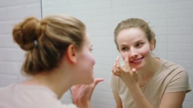 Sabah banyoda mutlu ve güzel bir kadın aynaya bakıyor. Güneşin gökyüzünü korumak için yüz kremi sürüyor.