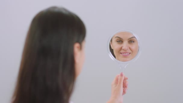 Adult woman very beautiful holding a makeup mirror on hand and smiling large she looking through the mirror straight to the camera — Stock Video