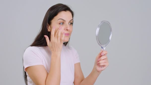 Hermosa mujer adulta con la piel perfecta y sonrisa sosteniendo un espejo que mira a su cara en el concepto de fondo blanco de cosméticos tratamiento de la piel — Vídeo de stock