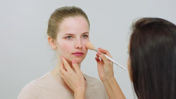 V přední části fotoaparátu na bílém pozadí make-up umělkyně vzít její štětec a aplikovat nějaké make-up produkty na modelu krásy dáma s perfektní pletí. 4k — Stock video