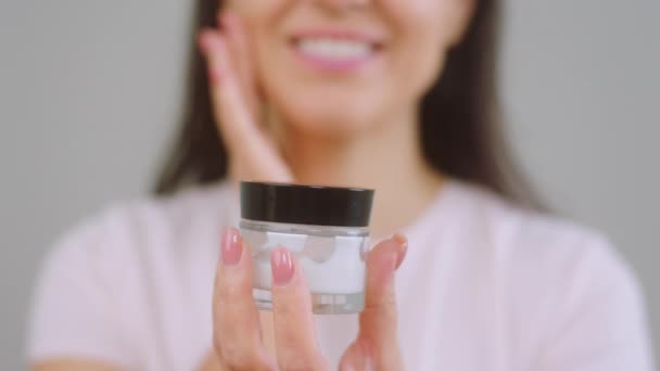 Részletek videó rögzítése közelkép nő gazdaság hidratáló krém a háttérben ő megérinti az arcát, és mosolygós nagy koncepció kozmetikumok bőrápoló kezelés — Stock videók