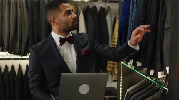 Egy luxus öltönyüzletben afro-amerikai tervező használja a laptopot, hogy ellenőrizze az újonnan érkezett öltöny kollekciót, és tegyen néhány értesítést a laptopon — Stock videók