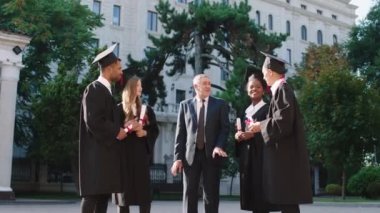 Mezuniyet günündeki mutlu bir grup öğrenci, üniversite profesörleriyle birlikte diplomalarını üniversite bahçesinde tutarak heyecanlı bir konuşma yapıyorlar.