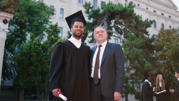 Bien parecido profesor universitario y estudiante graduado multirracial posando delante de la cámara con diploma muy emocionado — Vídeos de Stock