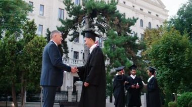Mezuniyet gününde iyi görünümlü bir adam üniversite müdürüne öğrencilerden birini verdi sonra da diplomayla birlikte kameranın önünde poz verdi.