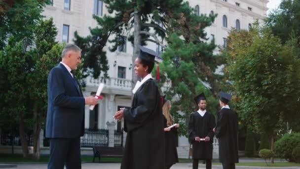 Señora negra graduada feliz tomando su diploma del profesor de la universidad en el jardín de la universidad delante de la cámara — Vídeos de Stock