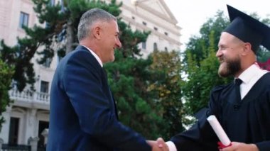 Mezuniyet gününde heyecanlı bir adam üniversitenin bahçesinde diplomasını aldıktan sonra babasıyla kucaklaşıyor.