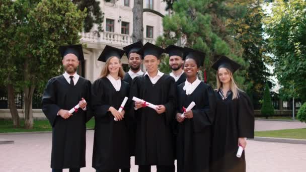Группа мультирасовых выпускников, позирующих счастливыми перед камерой в день окончания школы, с дипломами, которые они позируют в парке колледжа — стоковое видео