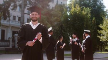 Üniversite bahçesinde mezuniyet günü kameranın önünde mezuniyet şapkası takmış kameraya bakarak gülümseyen, heyecanlı, büyük mezun adam. 4k