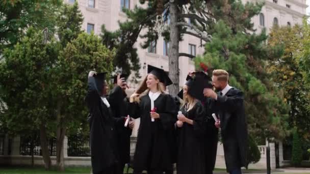 Grupo multiétnico de estudantes graduados após a formatura no parque universitário vomitou as tampas de formatura muito animado e segurando os diplomas — Vídeo de Stock