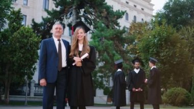 Heyecanlı, harika görünümlü, diplomalı bir bayan. Babası ile birlikte kameranın önünde poz veriyor. Çok heyecanlı, gülümsüyor.