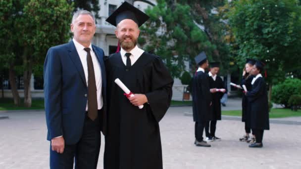 Día emocional para el padre y el hijo en la graduación que posan juntos delante de la cámara mientras sostienen el diploma — Vídeos de Stock