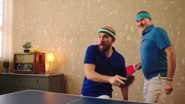 Heyecanlı ve gülümseyen iki erkek grubu. Biri genç, biri yaşlı, birlikte masa tenisi oynuyorlar. Kazanırlar ve mutlu bir şekilde sarılıp sırıtırlar. 4k