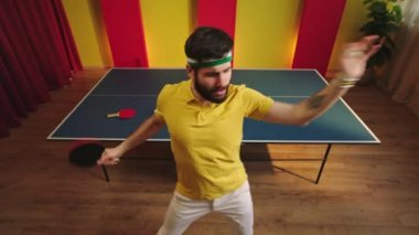 Komik ve çılgın masa tenisi oyuncusu, masa tenisi oyununu kazandıktan sonra kameranın önünde heyecan içinde dans ediyor. Raket ve topla oynuyor.