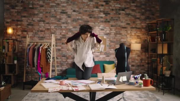 Mutlu ve çılgın moda tasarımcısı bayan çalışma masasının üzerinde kırmızı bir elbiseyle dans ediyor ve terzi atölyesinde eğleniyor. — Stok video