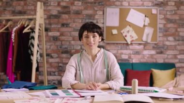 Küçük işletme atölyesi moda tasarımcısı güzel kıyafet tasarımcısı kadın iş yerinde otururken kameranın önünde poz veriyor ve bazı çizimler yapıyor.