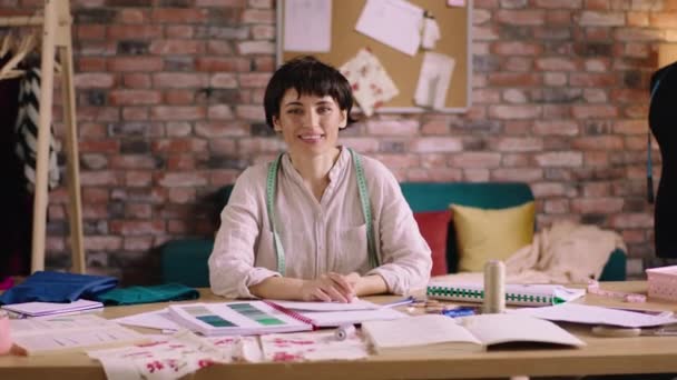 Dobrze wyglądająca projektantka mody w swoim biurze mody pozowała przed kamerą uśmiechając się uroczo, siedząc na stole w miejscu pracy i trzymając ołówek w dłoni. Strzał na ARRI Alexa Mini — Wideo stockowe