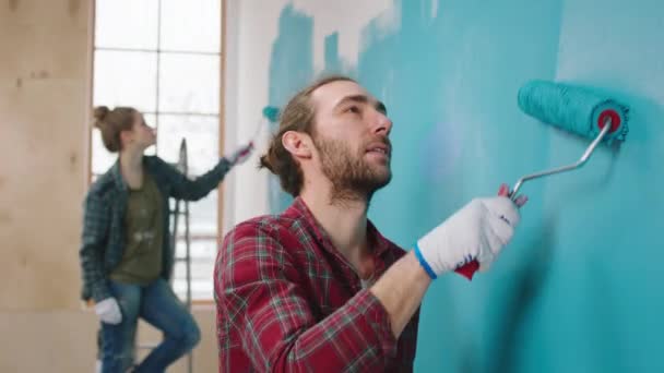 A kamera előtt jóképű férfi és a felesége együtt festik a falakat a nappaliban kék színben a fali kefe görgővel.. — Stock videók