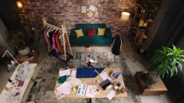 Üst Manzara. Bir adam çok rahat ve iyi dekore edilmiş bir ev ofisinde, masasında otururken bir uçakla oynuyor ve mavi kumaş parçasıyla tasarımına devam ediyor.