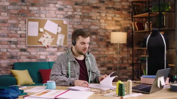 In front of the camera at tailoring atelier good looking man tailor speaks using the headphones with some customer he take the online order — Stock Video