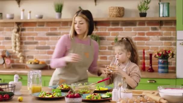 In de keuken met een rustiek design voegen de moeder en haar schattige dochter die samen het ontbijt bereiden wat vers sap in het glas toe aan haar dochter op het keukeneiland. — Stockvideo