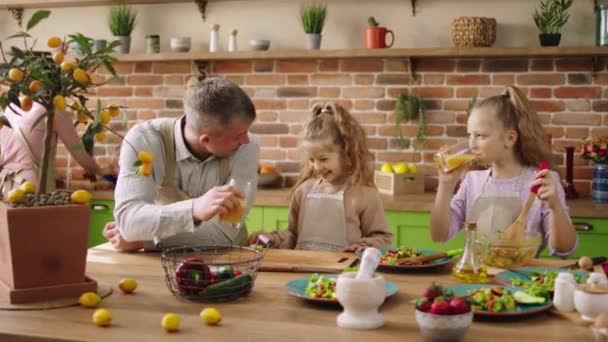 На современном деревенском острове на кухне харизматичный папа вместе с дочерьми готовит завтрак. Он дает дочери свежий апельсиновый сок. — стоковое видео