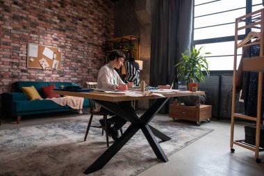 Terzi atölyesinde güzel bir kadın tasarımı çizimlerini günlükten analiz ediyor ve ofisinde yaratıcı bir gülümsemeye sahip.