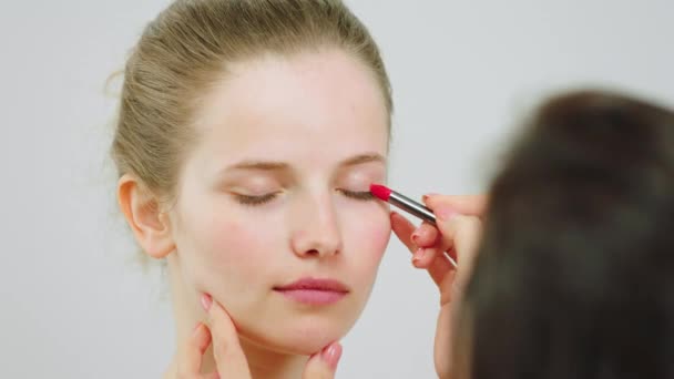 Λήψη βίντεο closeup μιας κυρίας μοντέλο με τέλειο δέρμα στο λευκό φόντο στέκεται, ενώ άλλες γυναίκες makeup artist κάνουν γυμνό μακιγιάζ χρησιμοποιώντας τα πινέλα μακιγιάζ — Αρχείο Βίντεο