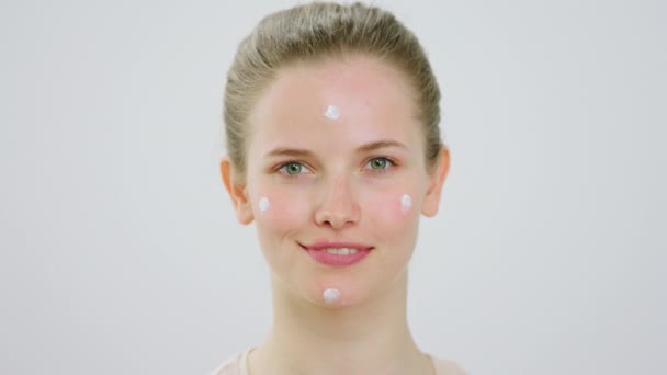 In front of the camera closeup taking portrait of young amazing looking Caucasian lady with perfect skin looking straight to the camera and smiling while have some moisturising skin cream on her face — Vídeos de Stock