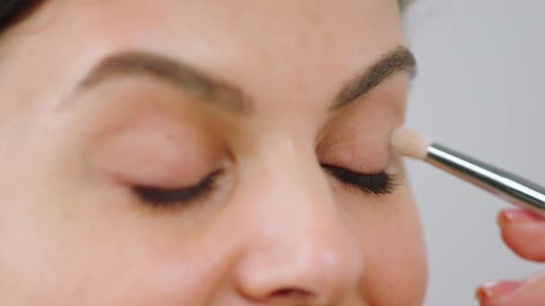 Gros plan sur la caméra sur fond blanc en prenant la vidéo d'une femme mature à la recherche étonnante avec une peau parfaite, elle applique des produits de maquillage sur ses yeux à l'aide d'un pinceau de maquillage — Video