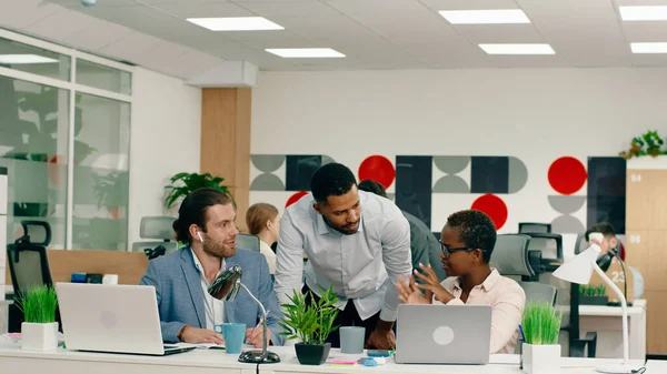 two African co- workers are talking about work eachother out in a large modern office, while a handsome man beside them does his work