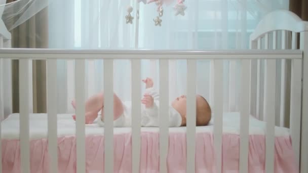 Pretty baby girl very handsome laying down on her baby cot alone playing with hands and legs excited she is concentrated at cot toys looking up — 비디오