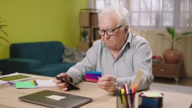 Charismatic old man at home using the smartphone to pay something online from the credit card he holding his credit card and type the card number