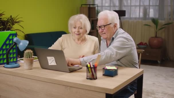 Hermosa pareja de ancianos en casa en la sala de estar se emocionan mientras ven algo en el portátil juntos en la sala de estar que sonríen grandes y abrazándose mutuamente — Vídeos de Stock