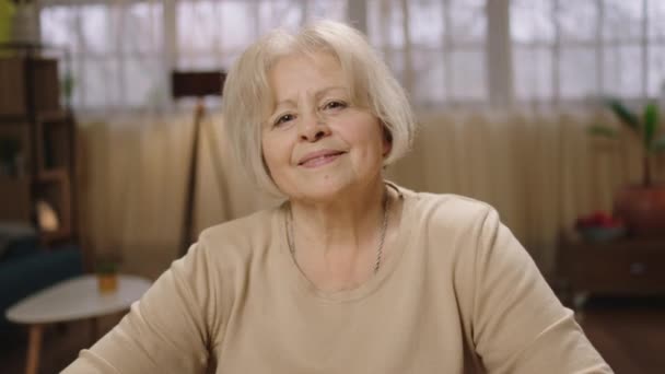 Titta rakt på kameran gör porträtt av en vacker gammal kvinna hemma hon ler söt — Stockvideo