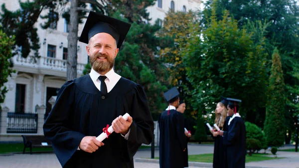 Uomo carismatico nel giardino del college in posa davanti alla telecamera dopo la laurea indossa un abito di laurea e cappello e in possesso di diploma — Foto Stock