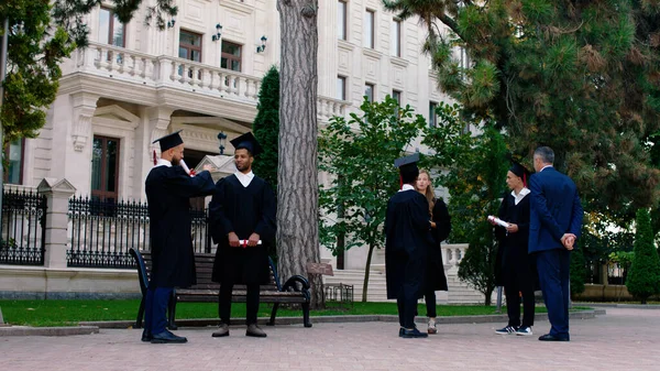 Mezuniyet gününde modern kolej parkında birkaç fotoğraf çekmek için bekleyen çok ırklı öğrencilerin mezuniyet kıyafetleri ve şapkaları içinde üniversite müdürüyle tartışmaları var.