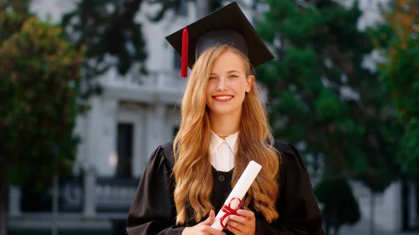 Hermosa con una gran sonrisa graduada posando frente a la cámara mientras sostiene su diploma en el jardín de la universidad —  Fotos de Stock