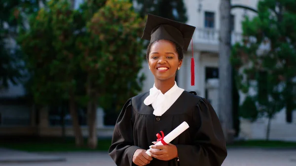 Beautiful black woman graduate posing with her diploma in front of the camera on the college garden she wearing graduation cap and suit — Foto Stock