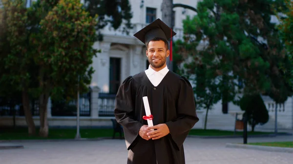 Bel ragazzo nel suo giorno di laurea indossa un abito e un cappello di laurea e posa davanti alla telecamera con il suo diploma nel giardino del college — Foto Stock