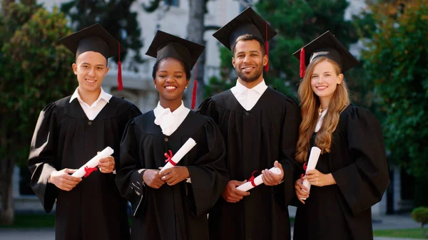 Gruppo di studenti multietnici si è laureato in posa davanti alla telecamera con i loro diplomi e sentirsi eccitati nel giardino del college indossando tuta di laurea e berretti — Foto Stock
