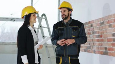 Kameranın önünde inşa halindeki modern bina. Güvenlik miğferi denetçisi olan güzel bir kadın inşaat işçisiyle konuşuyor. Son çalışma planını inceliyorlar.