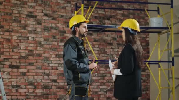 Attractive man supervisor at construction site discussing with the architect woman in a black suit about the plan of work they wearing safety equipment — Stok video