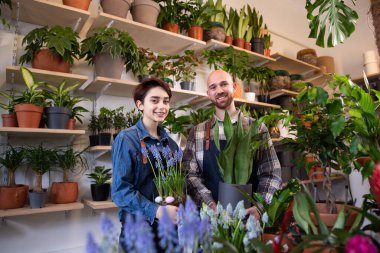 Çiçekçinin önünde çiçekçi adam ve çiçekçi kadın saksıyı incelerken sohbet ediyorlar.