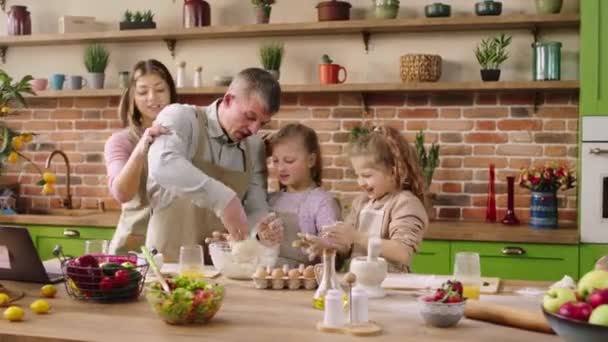 Na velkém kuchyňském ostrově šťastní nadšení rodiče spolu se svými dvěma dcerami připravují těsto na lahodný dezert táta tvrdé míchání těsta maminka a dcery mu pomáhají — Stock video