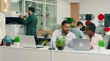 In a vivid office, two very attractive black assistants are sitting at their work tables and talking while a woman wearing a white blazers walks by