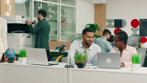In a vivid office, two very attractive black assistants are sitting at their work tables and talking while a woman wearing a white blazers walks by — Stok video