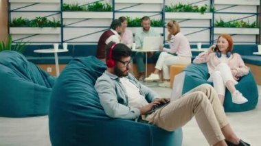 Çok yakışıklı genç bir adam laptopuyla çalışırken fasulye koltuğu üzerinde oturuyor, diğer işçiler arkada toplantı yapıyor ve kızıl saçlı bir kadın telefonunda.
